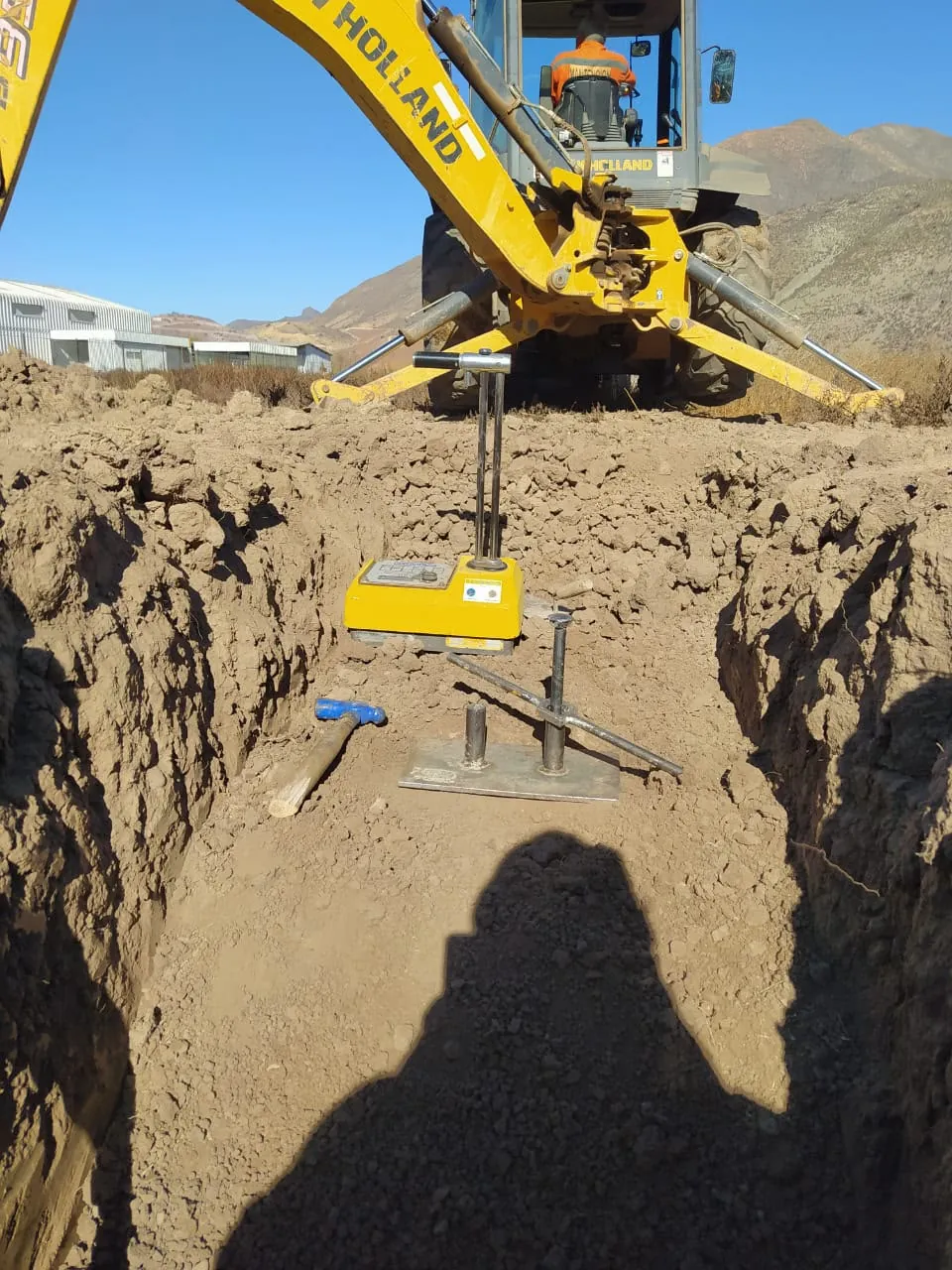 Densímetro nuclear amarillo en una calicata, con una retroexcavadora y los cerros al fondo