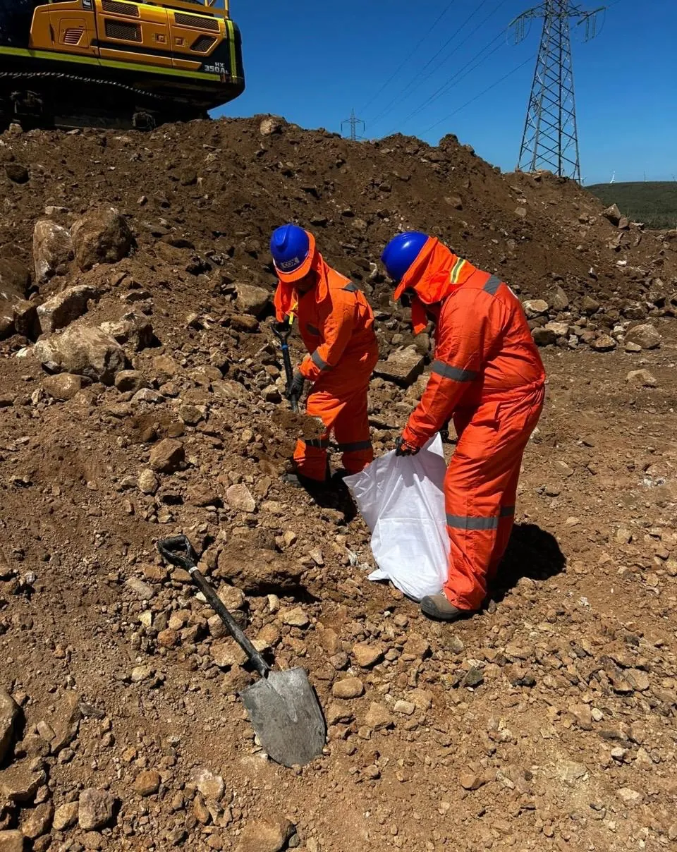 Dos técnicos llenando un saco con material de un acopio, con pala y maquinaria detrás